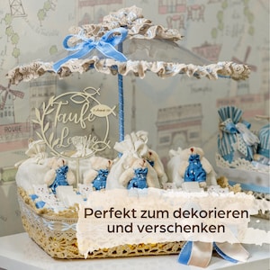 May include: A decorative basket with a clear umbrella-like top, decorated with blue ribbons and lace. Inside, small teddy bears in denim overalls are arranged around a wooden sign that reads "Taufte Leonie". The basket is decorated with white and blue accents.