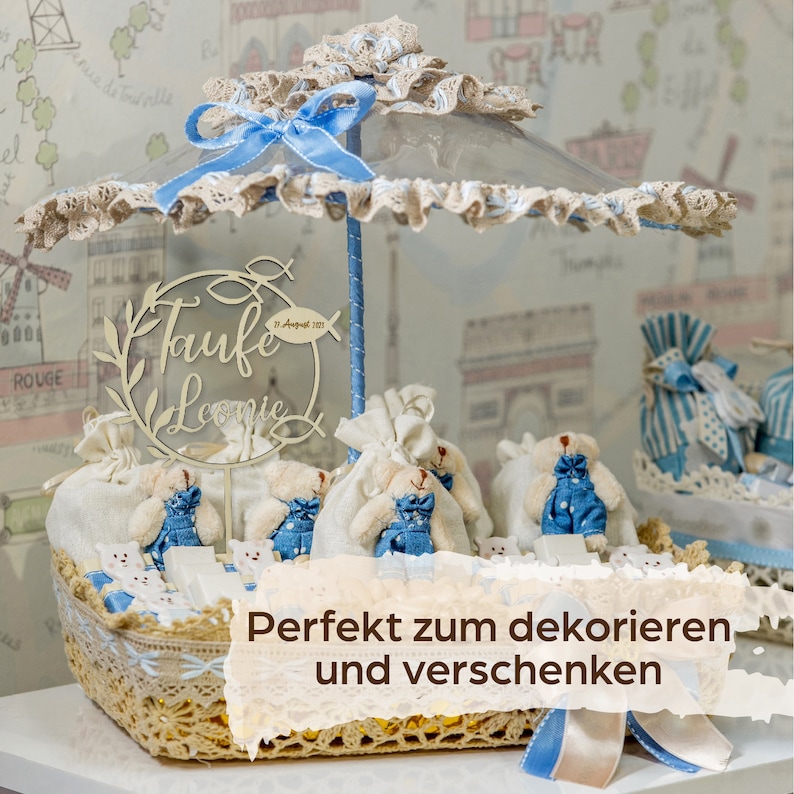 May include: A decorative basket with a clear umbrella-like top, decorated with blue ribbons and lace. Inside, small teddy bears in denim overalls are arranged around a wooden sign that reads "Taufte Leonie". The basket is decorated with white and blue accents.