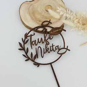 May include: A dark brown wooden cake topper with the words "Taufe Nikita" in cursive script, encircled by a wreath of leaves. The date "20. August 2023" is also included. The topper is designed for a baptism celebration.