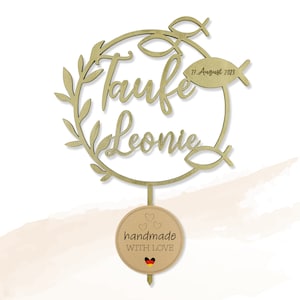 May include: A wooden cake topper with the words "Taufe Leonie" and the date "27. August 2023". The design includes a circular wreath, fish symbols, and a small tag that says "handmade WITH LOVE" with a German flag.