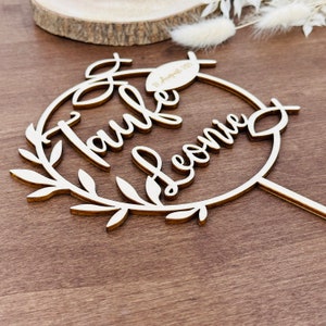 May include: A wooden cake topper with the words "Taufe Leonie" in cursive script, surrounded by a wreath of leaves. The date "11. August 2023" is also included. The topper is made of light-coloured wood.