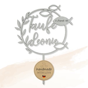 May include: A silver cake topper with the words "Taufe Leonie" and the date "27. August 2023". The design includes a circular wreath, fish symbols, and a small tag that reads "handmade WITH LOVE".