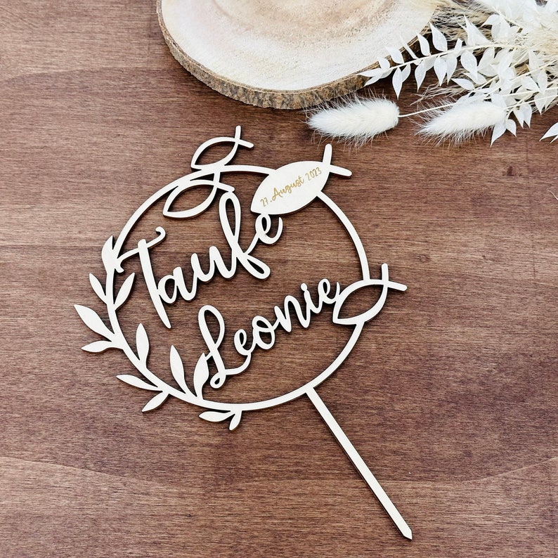 May include: A white wooden cake topper with the words "Taufe Leonie" in cursive script, encircled by a wreath design. A small fish shape and the date "21. August 2023" are also included. The topper has a long, thin stick for insertion.