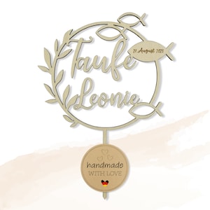 May include: A wooden cake topper with the words "Taufe Leonie" in script, encircled by leaves and fish motifs. The date "27. August 2023" is visible. A round tag at the base says "handmade WITH LOVE" and features a German flag.
