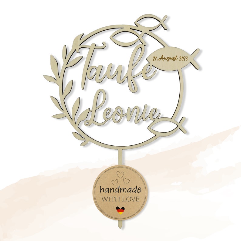 May include: A wooden cake topper with the words "Taufe Leonie" in script, encircled by leaves and fish motifs. The date "27. August 2023" is visible. A round tag at the base says "handmade WITH LOVE" and features a German flag.