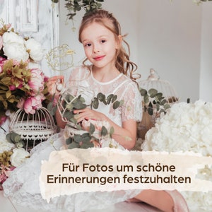 May include: A girl in a white lace dress smiles, surrounded by flowers and decorative cages. She holds a gold cake topper with the words "Thankful Blessings". The image includes the text "F&uuml;r Fotos um sch&ouml;ne Erinnerungen festzuhalten".