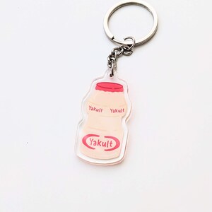 May include: A clear acrylic keychain shaped like a Yakult bottle with the Yakult logo printed in red on the front and back. The keychain is attached to a silver metal ring.