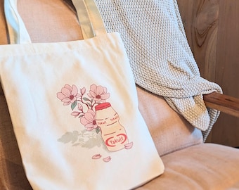 Puede incluir: Una bolsa de tela blanca con un estampado gráfico de una pequeña botella de Yakult rodeada de flores de magnolia rosas. El diseño incluye el texto del logotipo rojo de Yakult en la botella. La bolsa tiene dos asas largas para el hombro.
