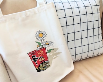Puede incluir: Un bolso de tela color crema con un estampado gráfico de un recipiente rojo de fideos instantáneos Shin Cup con flores de margarita blancas que emergen de la parte superior. El diseño incluye el carácter coreano negro para picante.