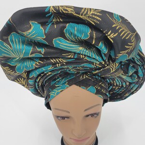 May include: A black head wrap with a teal and gold floral pattern. The fabric is folded and wrapped to create a turban style. The head wrap is on a mannequin head.