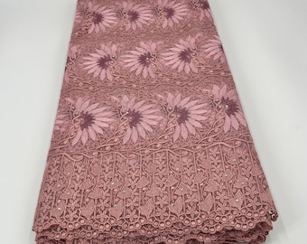 May include: A folded piece of dusty rose lace fabric. The fabric features a repeating floral pattern with detailed embroidery. The bottom edge has a scalloped design. The lace is likely used for clothing or decorative purposes.