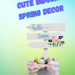 May include: A white wooden sign with the words "hello spring" and a floral design. The sign is attached to a small white pot with a pink and white gingham bow. The pot is filled with green grass and speckled Easter eggs.