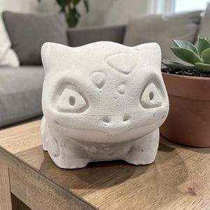 May include: A light gray concrete planter shaped like a Bulbasaur character. The planter has large eyes, a smiling mouth, and a textured surface. A small succulent plant in a brown pot is visible in the background.