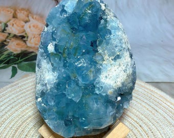 May include: A large, rough-cut celestite crystal geode in shades of light and medium blue. The crystal is mounted on a small wooden stand, with a woven mat and soft white background. The geode has a crystalline structure.