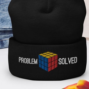 May include: Black beanie with embroidered text and a Rubik's Cube design. The text reads "PROBLEM SOLVED" in white. The Rubik's Cube is in its classic colors: red, blue, yellow, and orange. The beanie is made of a soft material.