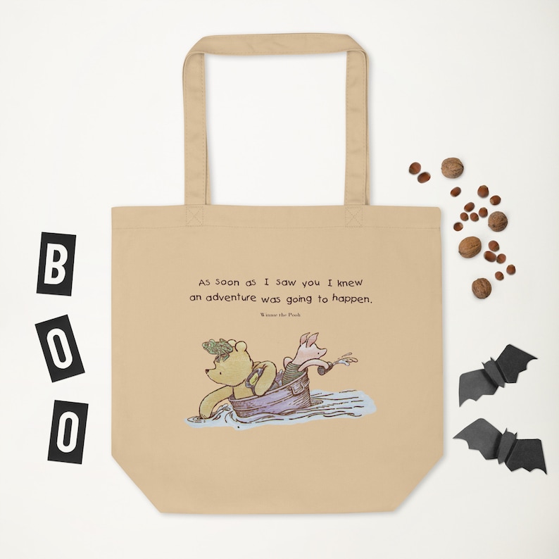 May include: A beige tote bag with a quote from Winnie the Pooh: "As soon as I saw you I knew an adventure was going to happen." The quote is in brown text and the image of Winnie the Pooh and Piglet in a boat is in the center of the bag.