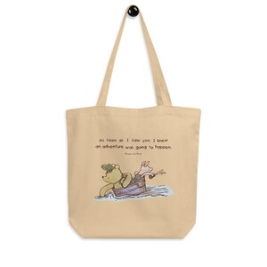 May include: A beige canvas tote bag with a Winnie the Pooh and Piglet illustration. The quote "As soon as I saw you I knew an adventure was going to happen" is printed on the bag.
