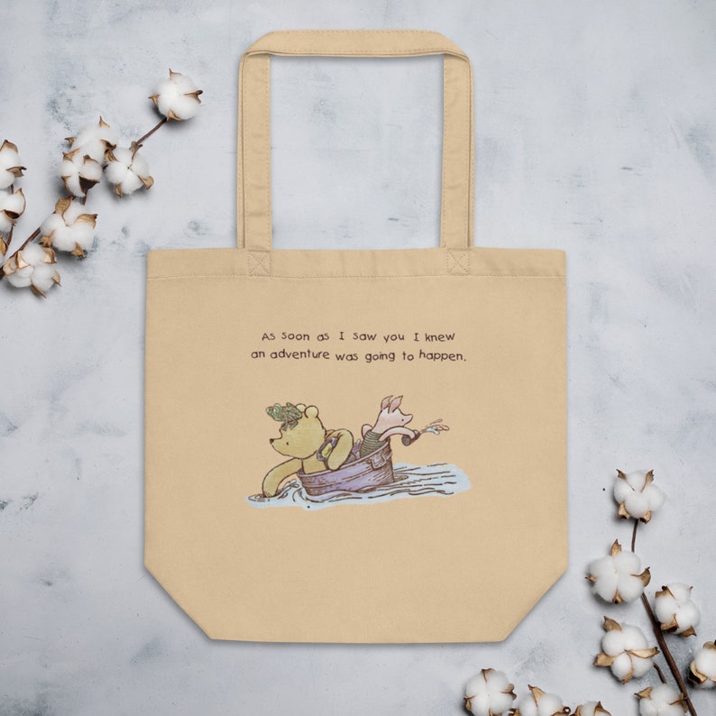 May include: A beige tote bag with a Winnie the Pooh and Piglet illustration. The text on the bag reads "As soon as I saw you I knew an adventure was going to happen."