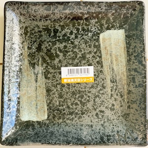 May include: A square, dark gray ceramic plate with a speckled glaze. The plate has two white brushstrokes on the surface. A yellow sticker with a barcode and the text "新海 天目シリーズ" is on the plate.