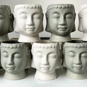 May include: A collection of ten small ceramic planters shaped like Buddha heads. The planters are in two colors: white and a light gray. Some of the planters have closed eyes, while others have open eyes.