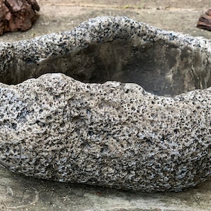 May include: A gray, textured, oval-shaped planter with a rough, uneven surface. The planter is made to look like a rock.