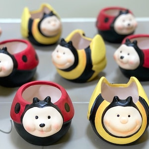 May include: A collection of ceramic planters shaped like cartoon bees and ladybugs. The planters are painted in bright colors, with yellow and black stripes for the bees and red with black spots for the ladybugs. Each planter has a smiling face.
