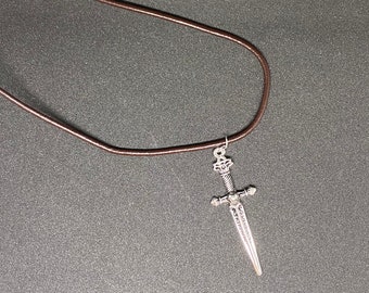 May include: A brown leather cord necklace with a silver sword pendant. The sword has a detailed design and a pointed tip.