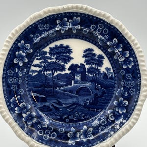 May include: A round ceramic plate with a scalloped edge featuring a white and deep blue transferware pattern. The center depicts a landscape with a stone bridge, trees, and buildings. The border is decorated with floral motifs and geometric patterns.