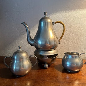 May include: A pewter tea set with a teapot, creamer, and sugar bowl. The teapot has a pear-shaped body and a brown handle. The creamer and sugar bowl are also pear-shaped.