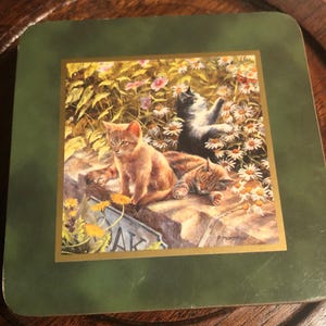 May include: A green coaster with a gold border featuring a painting of three kittens in a garden setting. The kittens are brown, black and white, and are playing amongst flowers.