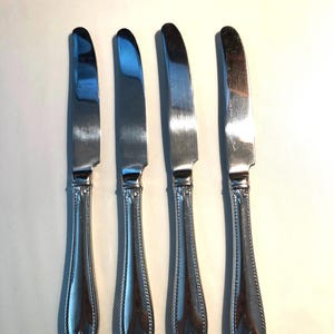 May include: Four silver dinner knives with ornate handles. The knives have a slightly curved blade and a rounded tip.