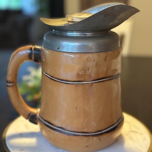 May include: A vintage pitcher with a bamboo-style design. The pitcher has a light brown body with dark brown accents and a silver metal lid and spout. The handle is also bamboo-style. The pitcher is sitting on a white marble-topped stand.