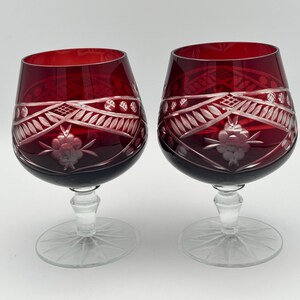 May include: Two ruby red crystal brandy snifters with intricate etched designs. The bowls are a deep red, transitioning to clear stems and bases. The etched patterns include floral and geometric elements, creating a vintage aesthetic.