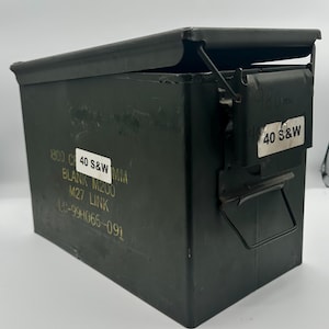 May include: An olive drab metal ammunition box with a hinged lid. The box has white labels with black text that reads "800 C 40 S&W MM BLANK M200 M27 LINK LC-99H066-091" and "40 S&W". The box is slightly open.