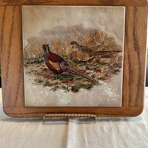 May include: A wooden framed tile with a painted image of two pheasants in a grassy field. The tile is on a gold stand.