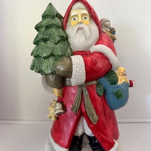 May include: A ceramic Santa Claus figurine in a red coat with white trim, holding a green Christmas tree and a blue sack filled with toys. The figurine has a long white beard and is standing on a snowy base.