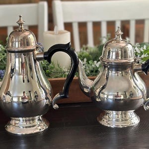 May include: Two silver teapots with black handles are displayed on a dark surface. The teapots have a shiny, reflective surface and a decorative finial on the lid. The teapots are part of a tea set.