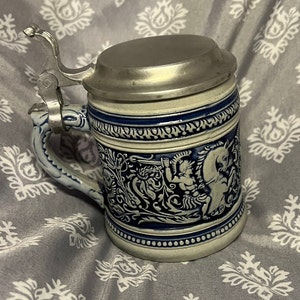 May include: A blue and white ceramic beer stein with a pewter lid. The stein features a detailed design of a knight on horseback.