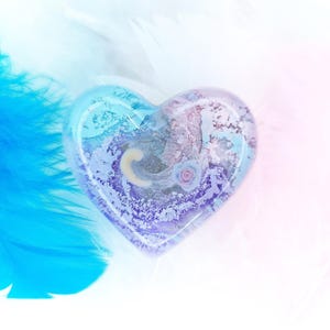 Könnte beinhalten: Ein herzförmiger Resin-Anhänger in Blau-, Lila- und Weißtönen. Er enthält eine kleine Rose und den Buchstaben "C". Der Hintergrund zeigt blaue Federn und einen sanften Farbverlauf.