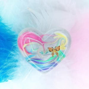 May include: A heart-shaped ornament with a vibrant, swirled pattern in pink, blue, yellow, and green. An orange butterfly is attached to the front. The item is set against a backdrop of white and blue feathers.