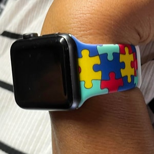 Puede incluir: Un Apple Watch negro con una banda de colores con estampado de piezas de rompecabezas. La banda est&aacute; hecha de silicona y tiene un respaldo blanco.