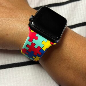 Puede incluir: Un Apple Watch negro con una banda de colores con estampado de piezas de rompecabezas. La banda es roja, azul, amarilla y verde.