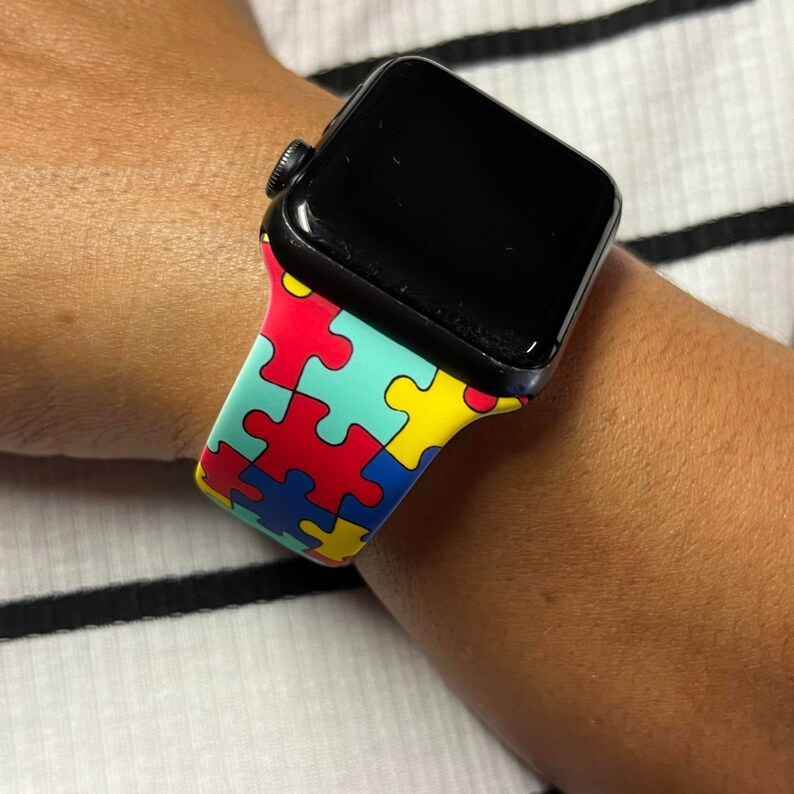 Puede incluir: Un Apple Watch negro con una banda de colores con estampado de piezas de rompecabezas. La banda es roja, azul, amarilla y verde.