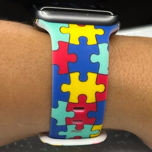 Puede incluir: Una banda de reloj colorida con estampado de piezas de rompecabezas en azul, amarillo, rojo y verde. La banda est&aacute; hecha de silicona y tiene una esfera de reloj negra.