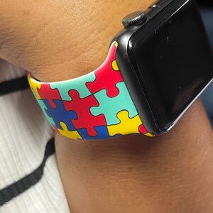 Puede incluir: Un Apple Watch negro con una banda de patr&oacute;n de piezas de rompecabezas blancas, azules, rojas y amarillas.