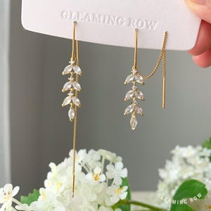 Può includere: Un paio di orecchini color oro con un design a forma di foglia. Ogni orecchino presenta una serie di pietre chiare e scintillanti, incastonate in una forma allungata e delicata. Il testo "GLEAMING ROW" è visibile.