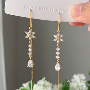 May include: A pair of gold-colored dangle earrings. Each earring features a floral design at the top, a series of clear, round stones, and a teardrop-shaped stone at the bottom. The earrings are suspended from a thin gold wire.