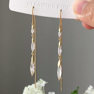 May include: A pair of gold-colored drop earrings with clear, marquise-cut crystals and small round crystals. The earrings have a delicate chain and a long, straight post. The brand name "GLEAMING ROW" is visible on the top.