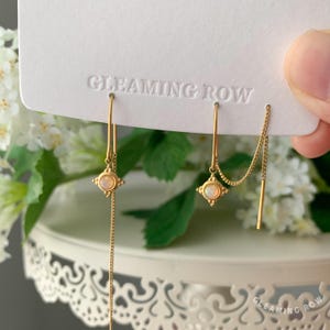 May include: Gold-toned earrings with a delicate design. Each earring features a small, square-shaped charm with a central stone and a dangling chain. The earrings are displayed on a white card with the text "GLEAMING ROW".