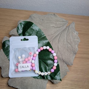 Könnte beinhalten: Ein handgefertigtes Armband mit dem Namen "CALLA" in goldenen Buchstaben. Das Armband hat rosa, weiße und lila Perlen. Ein kleiner Beutel mit passenden Perlen und Faden ist ebenfalls enthalten. Das Armband wird auf einem Blatt präsentiert.